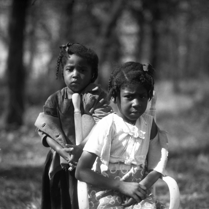 Two young girls look into the camera. One of them is seated in a small chair in a field. The other is standing behind the chair.