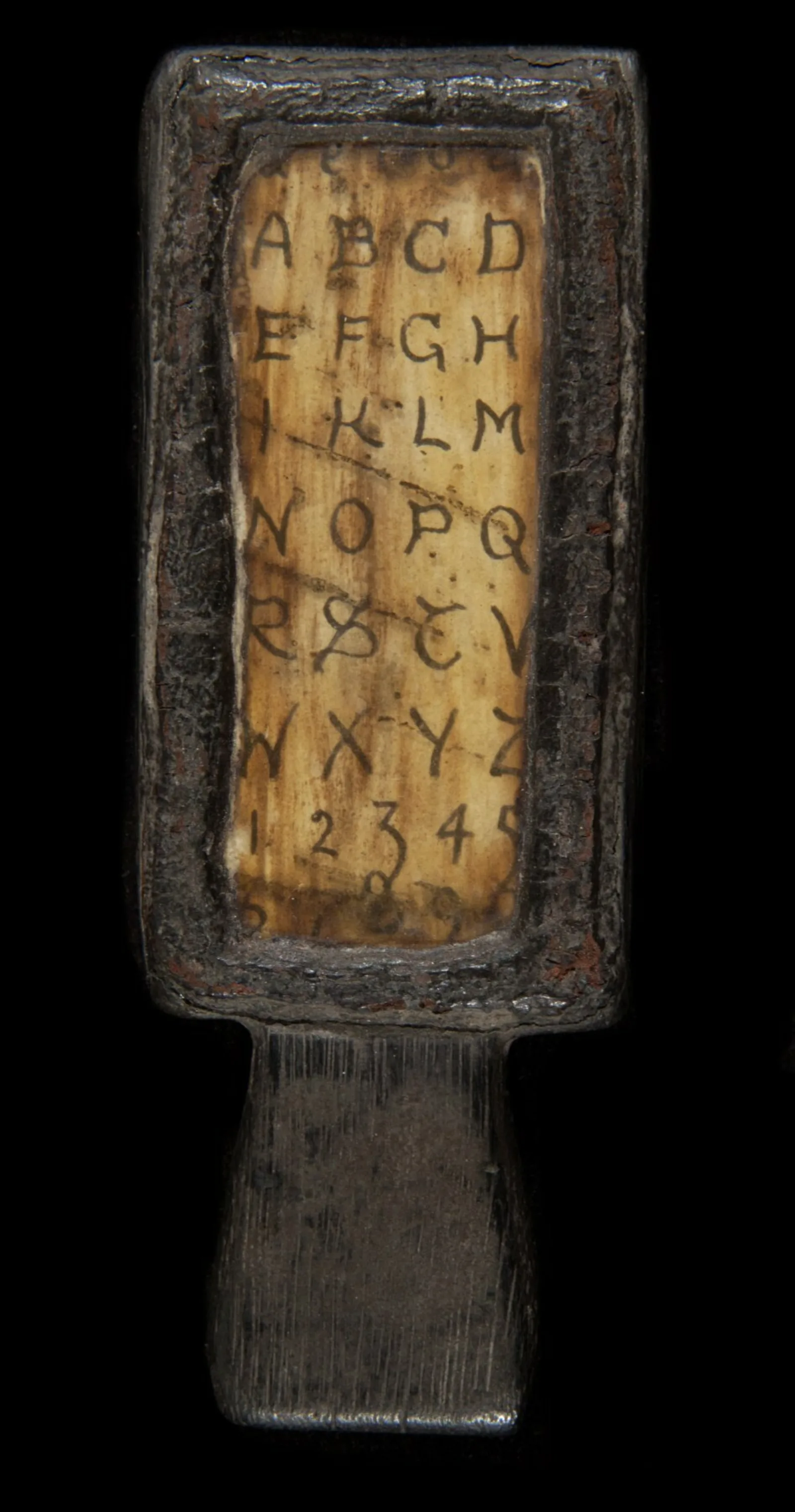 A small wooden paddle has a transparent screen running vertically. Under the screen is a handwritten alphabet. 