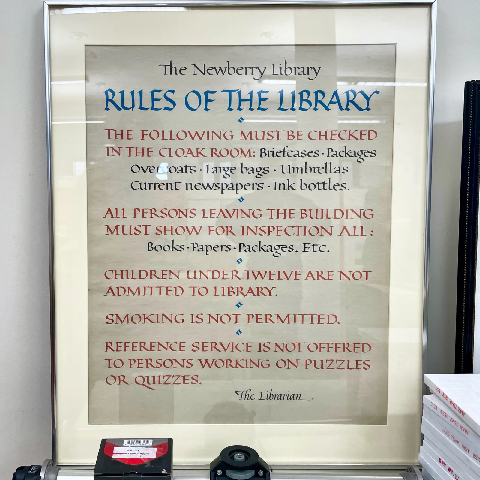 The sign reads the following: "The Newberry Library. Rules of the Library. The following must be checked at the cloak room: briefcases, packages, overcoats, large bags, umbrellas, current newspapers, ink bottles. All persons leaving the building must show for inspection all: books, papers, packages, etc. Children under twelve are not admitted to library. Smoking is not permitted. Reference service is not offered to persons working on puzzles or quizzes. The librarian."