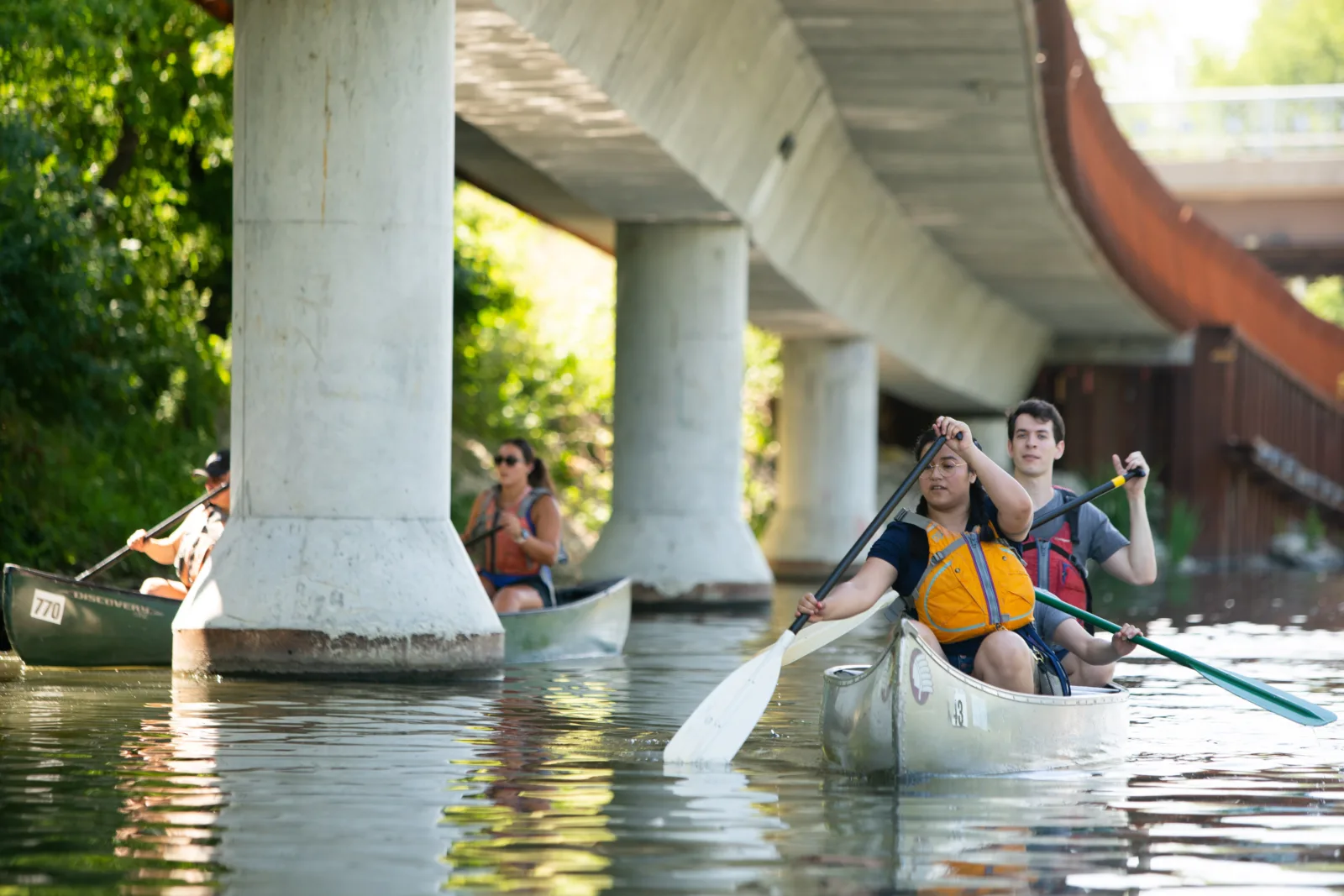 NCAIS participants paddle down the Chicago River. Behind them are large concrete poles and highways characteristic of the city's urban landscape.