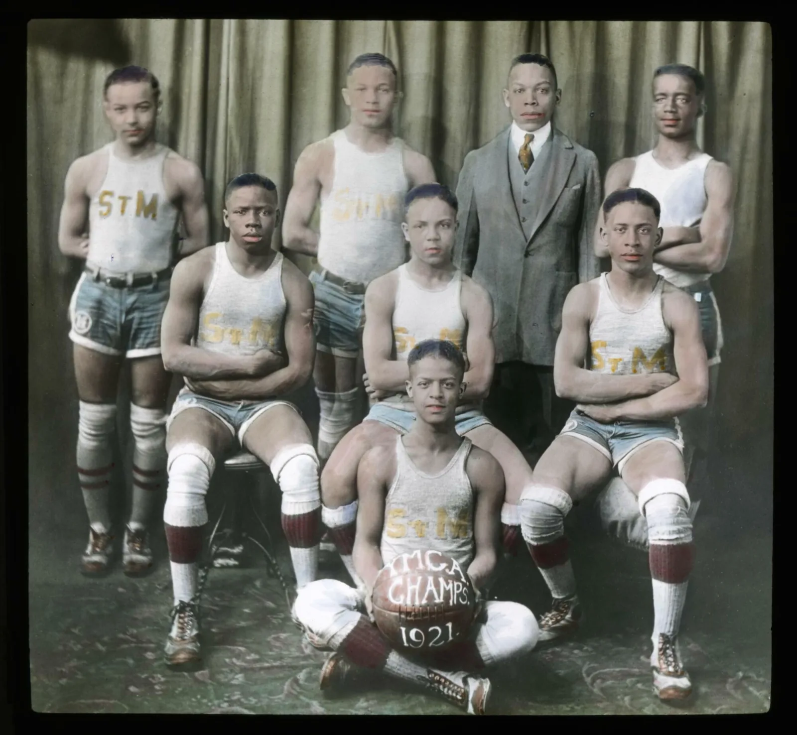 Youth basketball players sit or stand in three rows. They are wearing tank tops, shorts, long socks, and knee pads. The player sitting in the middle holds a basketball. On the basketball are the words “YMCA Champs 1921.”
