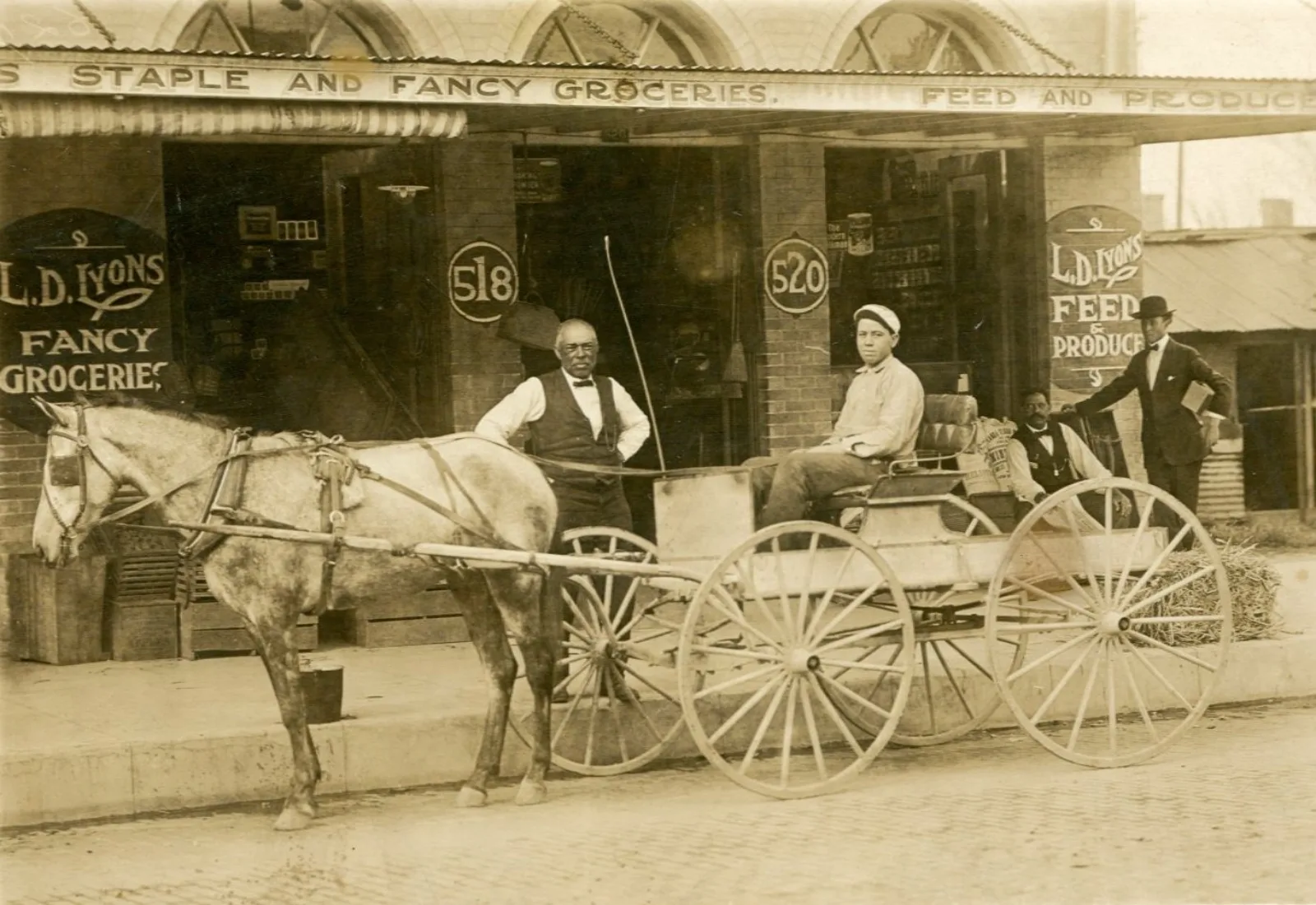 People pose around a horse and buggy. They're standing in front of a storefront with the words  "LD Lyons Fancy Groceries" on the facade.