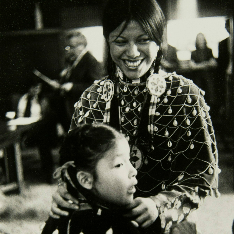 Barbara Bearskin with her hands on Kathy White's shoulders. Both are wearing their powwow regalia. D'Arcy McNickle is in the background of the photo.